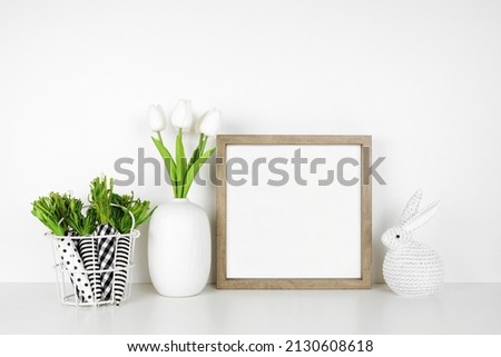 Mock up wood frame with Easter decor on a white shelf. Cloth carrots, knit bunny and tulip flowers. Square frame against a white wall. Copy space.