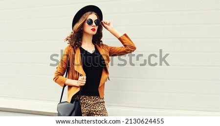 Beautiful stylish woman model wearing black round hat, brown jacket and handbag on gray background