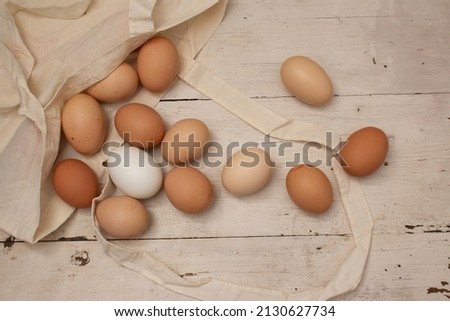 Top view. Food photography for holiday Happy Easter. Beige, white and chicken chicken eggs spilled out of canvas shopper bag on wooden vintage white table with scuffs and cracks. Village kitchen. Wood