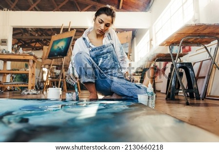Talented female artist making a painting on a canvas in her art studio. Skilful young painter squatting on the floor while working on a new artwork for her creative project. Royalty-Free Stock Photo #2130660218
