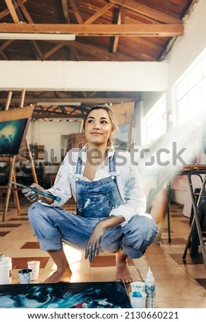 Pensive young artist squatting on the floor in her studio. Female painter contemplating new creative ideas for her art project. Imaginative young woman making a painting on a large canvas. Royalty-Free Stock Photo #2130660221