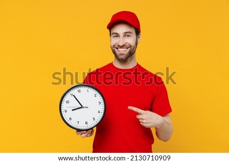Professional delivery guy employee man 20s in red cap T-shirt uniform workwear work as dealer courier hold in hands point finger on clock isolated on plain yellow background studio Service concept.