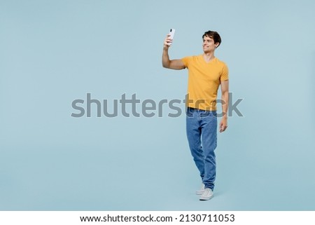 Full body young man 20s wear yellow t-shirt doing selfie shot on mobile cell phone post photo on social network isolated on plain pastel light blue background studio portrait. People lifestyle concept