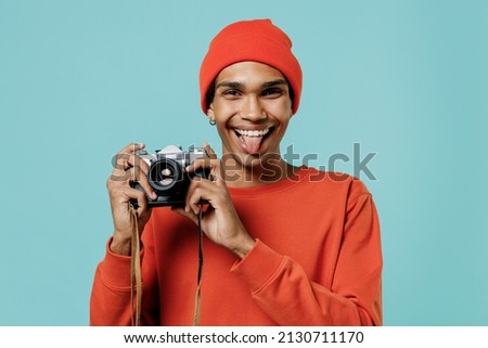 Young happy fun african american man in orange shirt hat taking photo picture on retro vintage photo camera show tongue isolated on plain pastel light blue background studio. People lifestyle concept Royalty-Free Stock Photo #2130711170