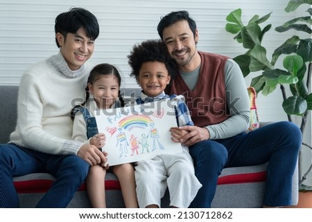 Happy Asian young LGBTQ gay couple with little cute adopted Caucasian and African kid smiling and showing rainbow family drawing in living room at home. LGBT diverse family concept