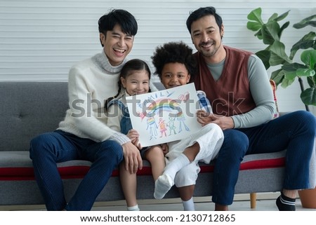 Happy Asian young LGBTQ gay couple with little cute adopted Caucasian and African kid smiling and showing rainbow family drawing in living room at home. LGBT diverse family concept