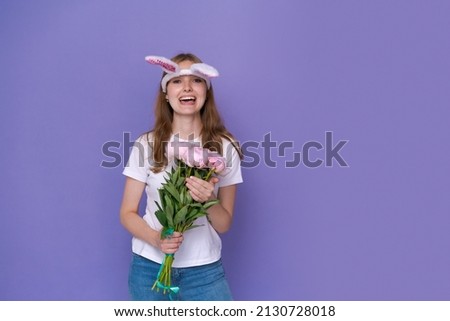 Easter holidays, spring concept - happy funny, crazy young girl in headband with rabbit ears holding a bouquet of pink peonies on a purple background. Banner, copy space for text, mockup