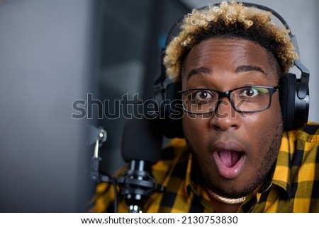 Portrait of an African radio host with glasses and with microphone, who opened his mouth in surprise and looks at the camera. Funny black man is delighted with what he heard in his headphones