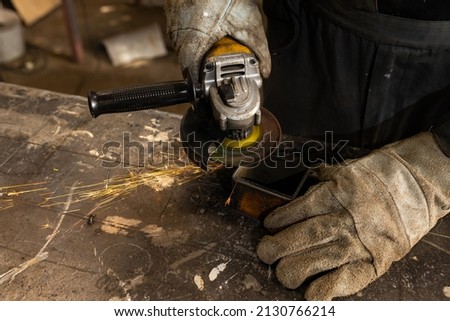 A man in overalls, gloves at the enterprise grinds metal products with an angle grinder on a metal workbench. Sparks fly.