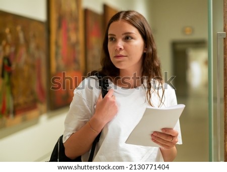 Portrait of a confident girl with an information booklet, standing in the hall of the historical museum next to the ..exhibit, located in a glass cabinet Royalty-Free Stock Photo #2130771404