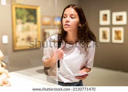 Attentive girl visitor looks with interest at the exhibit in the museum, located behind the glass Royalty-Free Stock Photo #2130771470