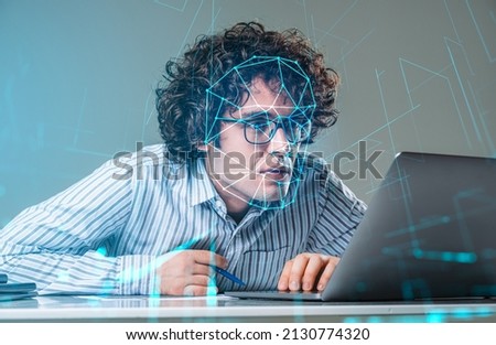 Young handsome curly businessman wearing formal shirt is working on laptop. Office workplace with desk and document in the background. Digital interface. Concept of facial recognition