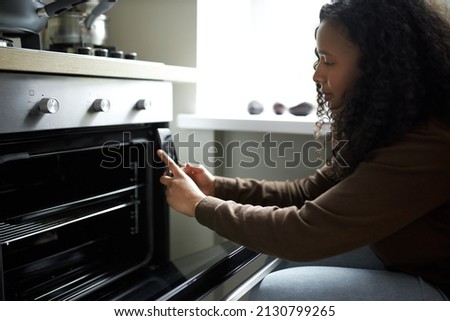 Side view of dark-skinned female blogger taking picture of her oven and empty tray, having vlog of cooking at social networks, creating new video of preparing delicious meal. Human and technology