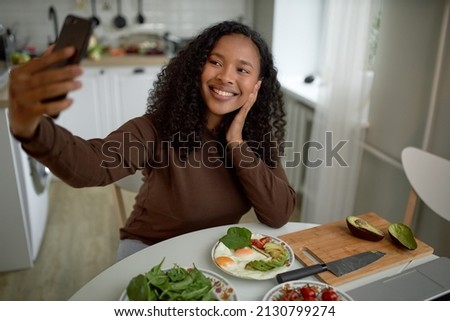 Sweet girl of 20s taking picture of herself having beautiful, colorful, healthy, tasty and rich breakfast containing fried eggs, avocado slices, spinach, sitting at table with knife and board