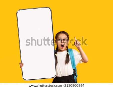 Wow, Great Offer. Excited Little Asian School Girl In Eyeglasses Holding Big Cell In Hand Pointing Finger Up Presenting Phone Empty Screen On Yellow Orange Studio Background. Smartphone Display Mockup