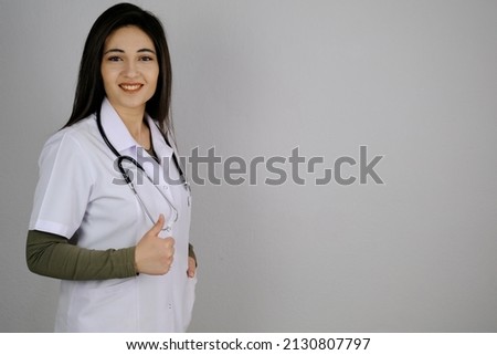 Positive Caucasian female doctor in lab coat showing thumb up gesture over grey studio background