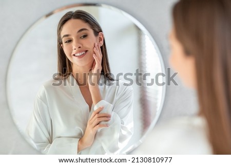 Self-Care Concept. Portrait Of Attractive Young Female Looking At Mirror, Beautiful Woman Wearing White Silk Robe Touching Soft Skin On Face And Smiling, Enjoying Her Reflection, Selective Focus Royalty-Free Stock Photo #2130810977