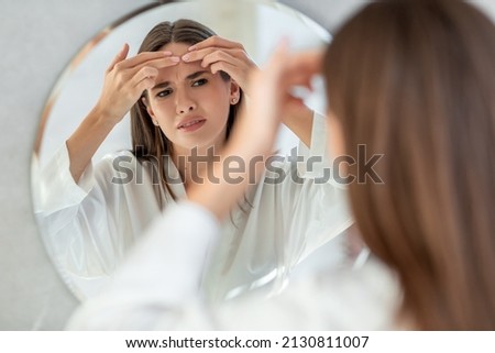 Problem Skin. Upset Young Female Popping Pimple On Forehead While Standing Near Mirror In Bathroom, Frowning Millennial Woman Inspecting Her Face, Suffering Acne, Selective Focus On Reflection Royalty-Free Stock Photo #2130811007