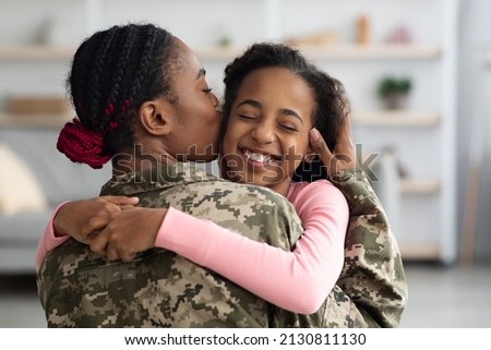 Loving black mother in military uniform kissing her happy daughter, african american female soldier return from army, hugging her child, home interior, closeup photo, copy space