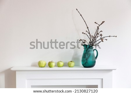 A green jug with cut poplar branches and four apples is on a white fireplace as a room decoration