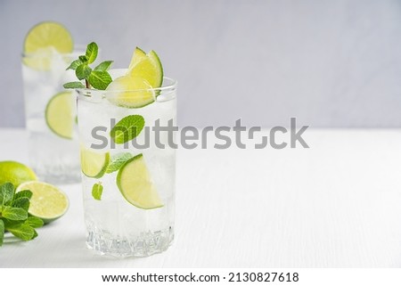 Organic cold refreshing lemonade drink or cocktail made of sparkling water, lime slices and fresh green mint leaves served in tall drinking glass with ice cubes on white wooden table with copy space Royalty-Free Stock Photo #2130827618