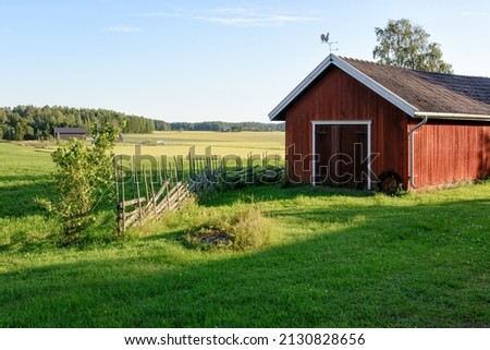 Traditional Finnish red wooden barn with roundpole fence and the weather vane on the roof. Old idyllic farm in the countryside. Royalty-Free Stock Photo #2130828656