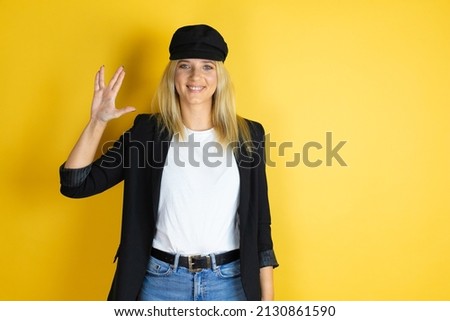 Beautiful woman wearing casual white t-shirt and a cap over isolated yellow background doing hand symbol