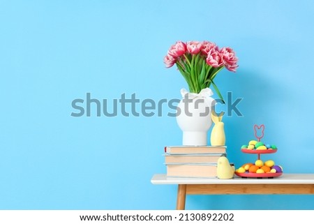 Vase with tulips, books, decor and Easter eggs on table near blue wall