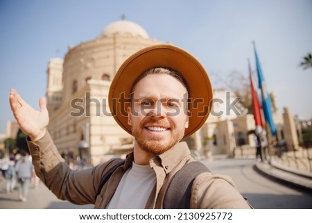 Young archaeologist man in hat takes selfie photo against background of ancient buildings. Concept archeological sit old city.