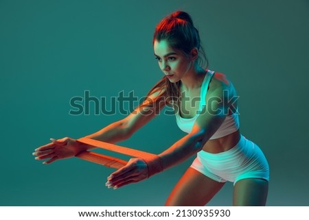 Portrait of sportive woman workout, doing exercises with sports equipment isolated on green studio background in neon light. Sport, gym, action, motion, beauty concept. Fitness, hobby, health Royalty-Free Stock Photo #2130935930