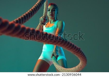 Ropes. Portrait of sportive woman workout, doing exercises with sports equipment isolated on green studio background in neon light. Sport, gym, action, motion, beauty concept. Fitness, hobby, health Royalty-Free Stock Photo #2130936044