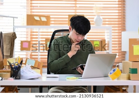 A portrait of an Asian man, e-commerce employee sitting in the office full of packages in the background using a laptop and writing a note, for SME business, e-commerce and delivery business.