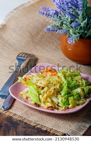 Eating cabbage fried or stewed with herbs, carrots, onions on a pink square plate, fork and knife on a burlap tablecloth on the table. Lavender flowers. Vegetarian food.