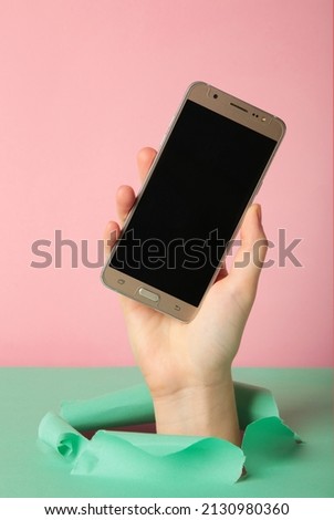 Closeup of female hand holding smartphone with white empty screen showing device close to camera breaking through pink paper sheet. Gadget display with free copy space, mockup. Top view