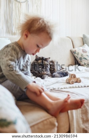 The kid, sitting on the couch, plays next to a resting pet cat