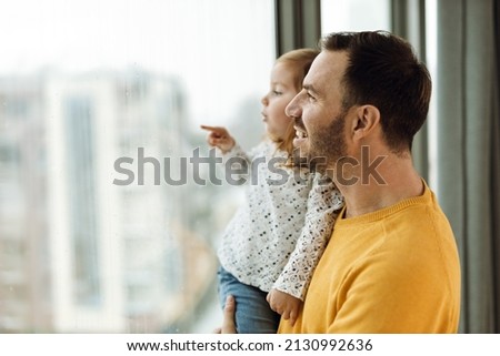 Happy father and his small girl looking through window from their home