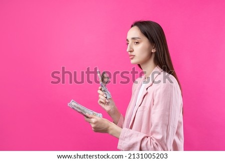 A woman in a pink jacket holds dollar banknotes, a puzzled business girl hides dollars standing on a red background.