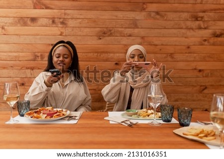 Two female friends taking pictures of their food in a restaurant. Two diverse young women having a wholesome lunch in a cafeteria. Millennial friends creating content for their social media blogs.