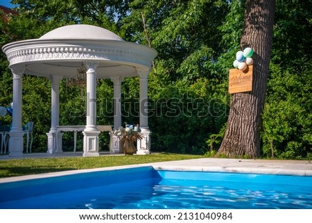 Luxury decoration setup by the pool for event celebration in backyard terrace with plaster pillars, with table, flowers, balloons and welcome table hanging on a tree. Natural background.