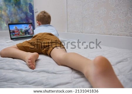 Barefoot child is watching entertainment content on a portable device while lying on a bed. A small blond boy in white wireless headphones uses a modern laptop on the bed.