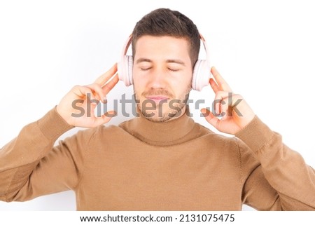 Pleased young caucasian man wearing turtleneck over white background enjoys listening pleasant melody keeps hands on stereo headphones closes eyes. Spending free time with music