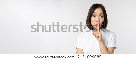 Close up portrait of young beautiful asian girl shushing, has secret, keep quiet silence gesture, press finger to lips, standing in tshirt over white background