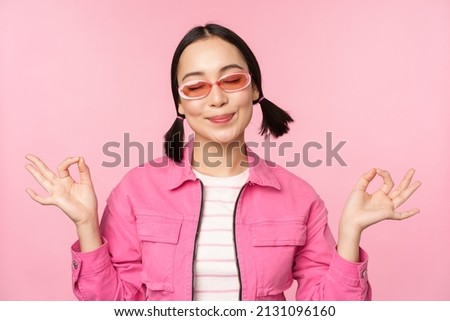 Mindfulness and wellness concept. Smiling korean girl in stylish outfit meditating, listening mantra, holds hands in zen, peace pose, practice yoga, standing over pink background