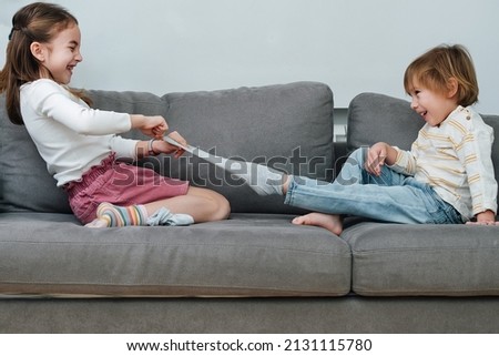 Laughing girl pulling off sock of a boy, they are preparing for a tickling competition. Sitting on a couch in a living room. Side view.