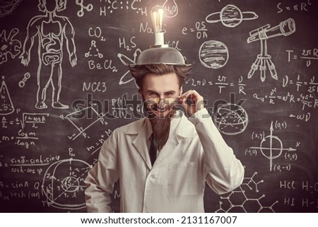 A smart eccentric scientist stands at the blackboard with scientific formulas and diagrams with a strange invention on his head and laughs madly. Science and new inventions. Royalty-Free Stock Photo #2131167007