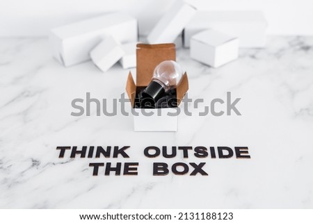 Think outside the box concept with idea lightbulb on top of white box and text on minimalist white marble background