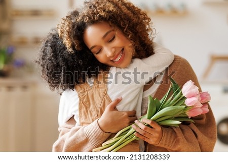 Little african american son congratulating mom with Mother's day at home and giving her fresh flower bouquet, happy mixed race woman mother embracing son. Family holidays and celebration concept Royalty-Free Stock Photo #2131208339