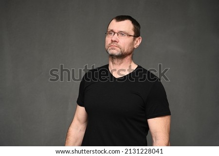 An adult man with glasses looks away. Portrait of a caucasian man in a black t-shirt on a gray background in the studio