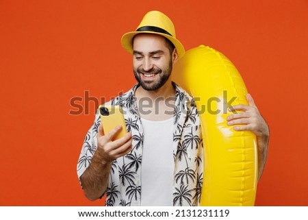 Young smiling happy tourist man in beach shirt hat hold inflatable ring hold in hand use mobile cell phone isolated on plain orange background studio portrait. Summer vacation sea rest sun tan concept