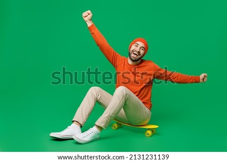Full body young smiling happy friendly fun cheerful cool man 20s wearing orange sweatshirt hat sit on skateboard with outstretched hands arms isolated on plain green color background studio portrait.